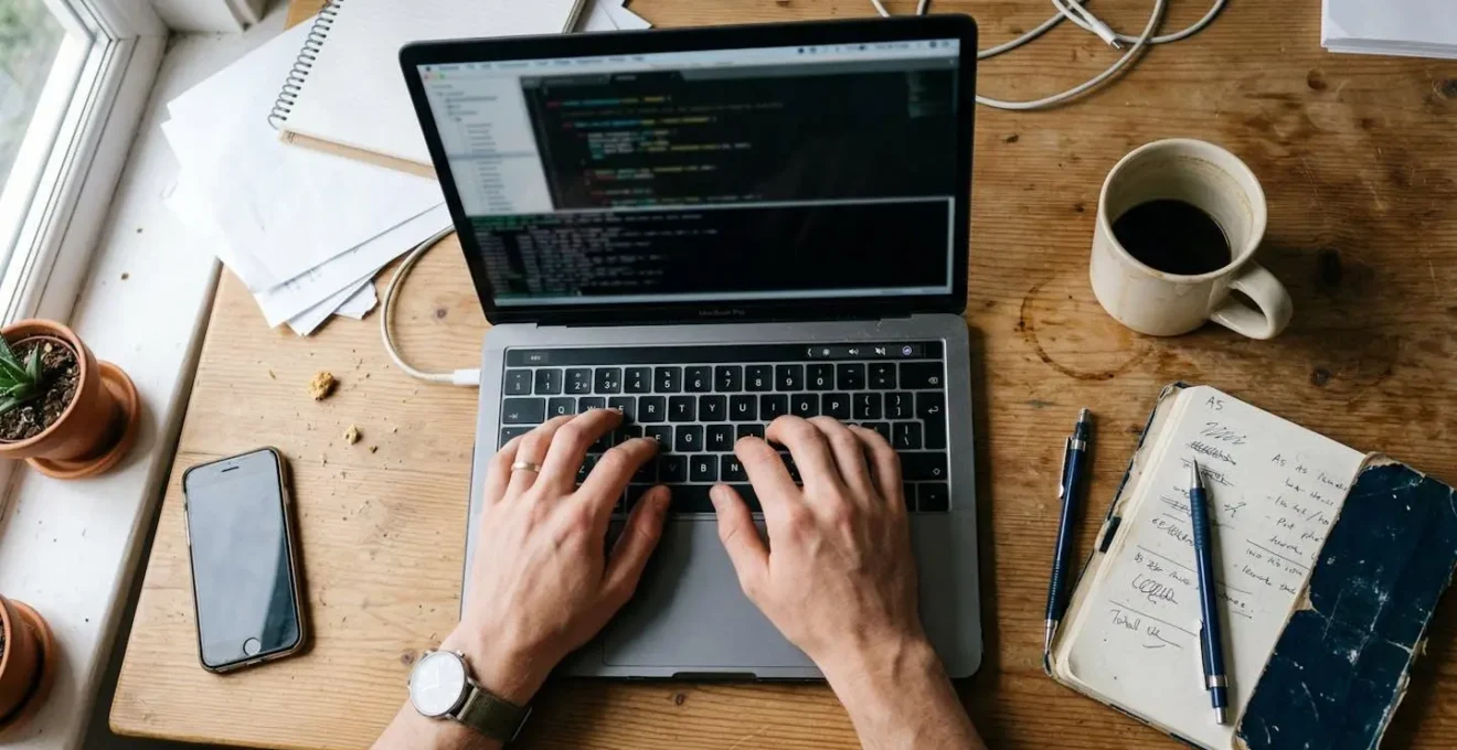 Bureau de travail vu de dessus avec ordinateur portable, tasse de café et carnet, mains posées sur le clavier
